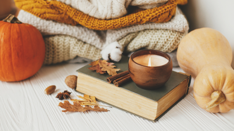 A closed book with a candle reseting on top, beside some sweaters and pumpkins